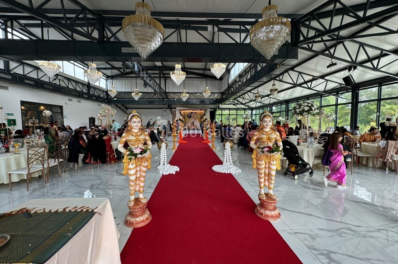 Salle de réception décorée pour un mariage avec statues traditionnelles, tapis rouge et invités assis à des tables.
