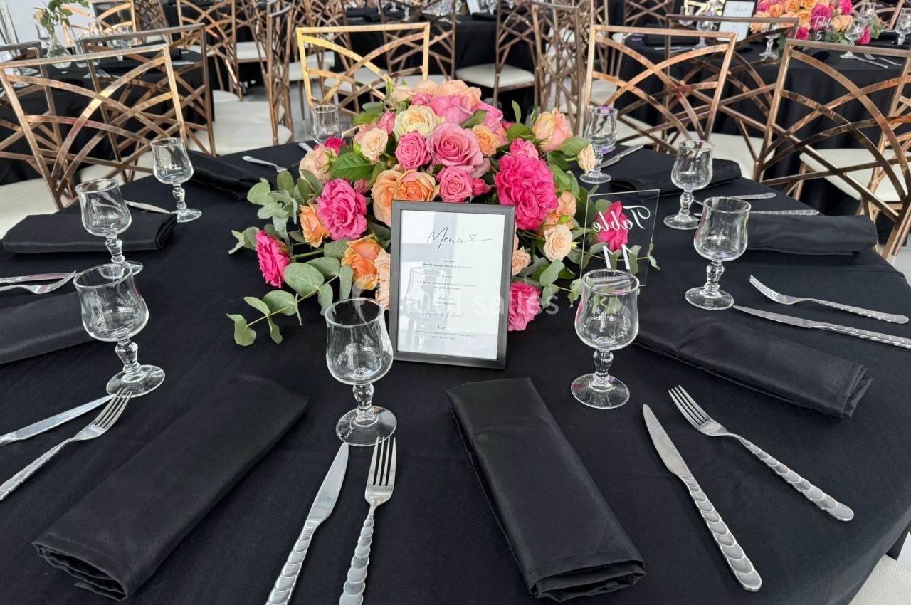 Table dressée avec une nappe noire, couverts argentés, verres, serviettes pliées et un centre de table floral coloré.