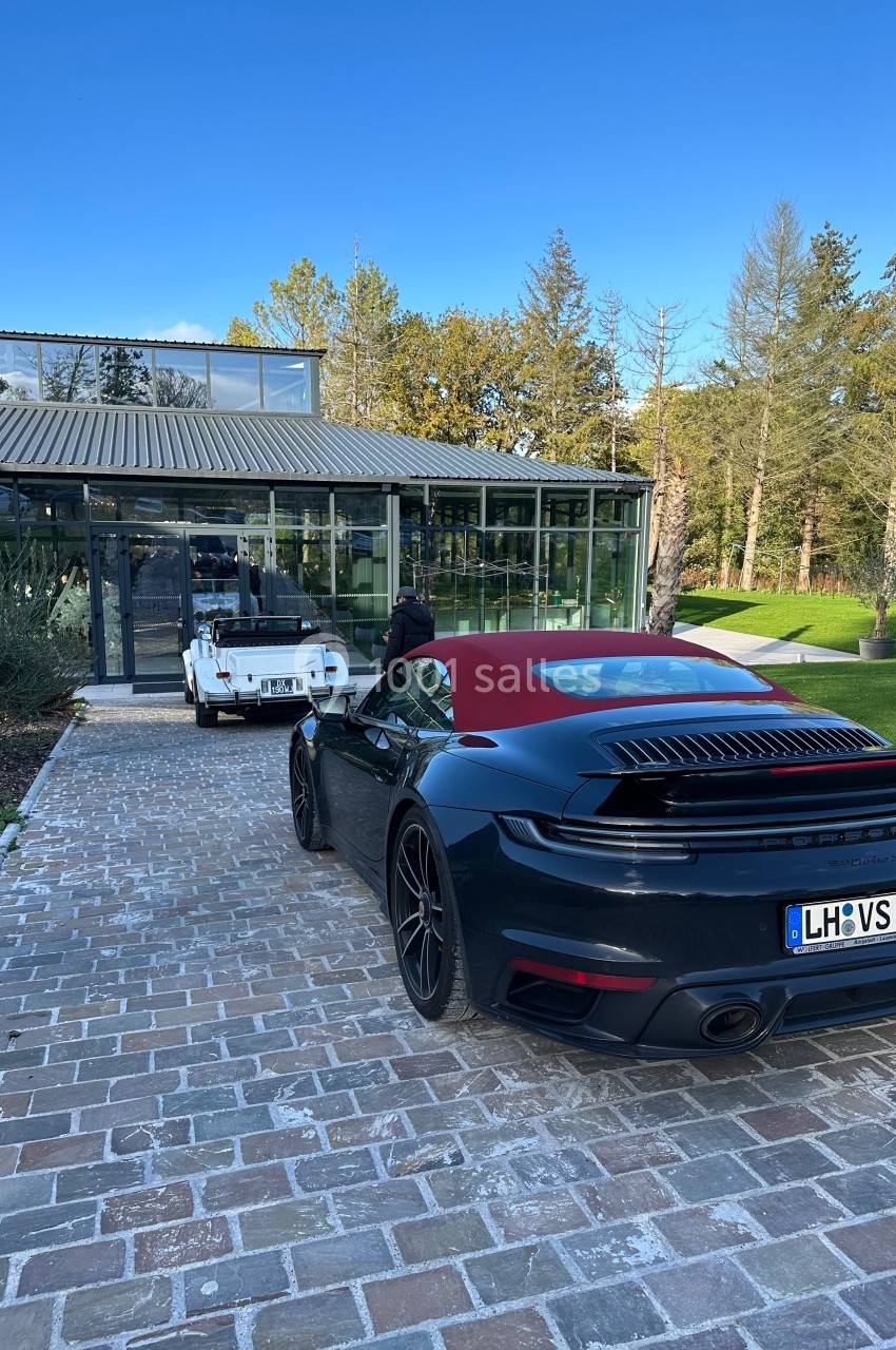 Voiture de sport noire garée sur une allée pavée devant une maison moderne en verre, avec un véhicule blanc à l'arrière.