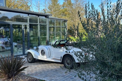 Voiture ancienne blanche stationnée devant une véranda moderne, entourée de végétation et d'arbres.