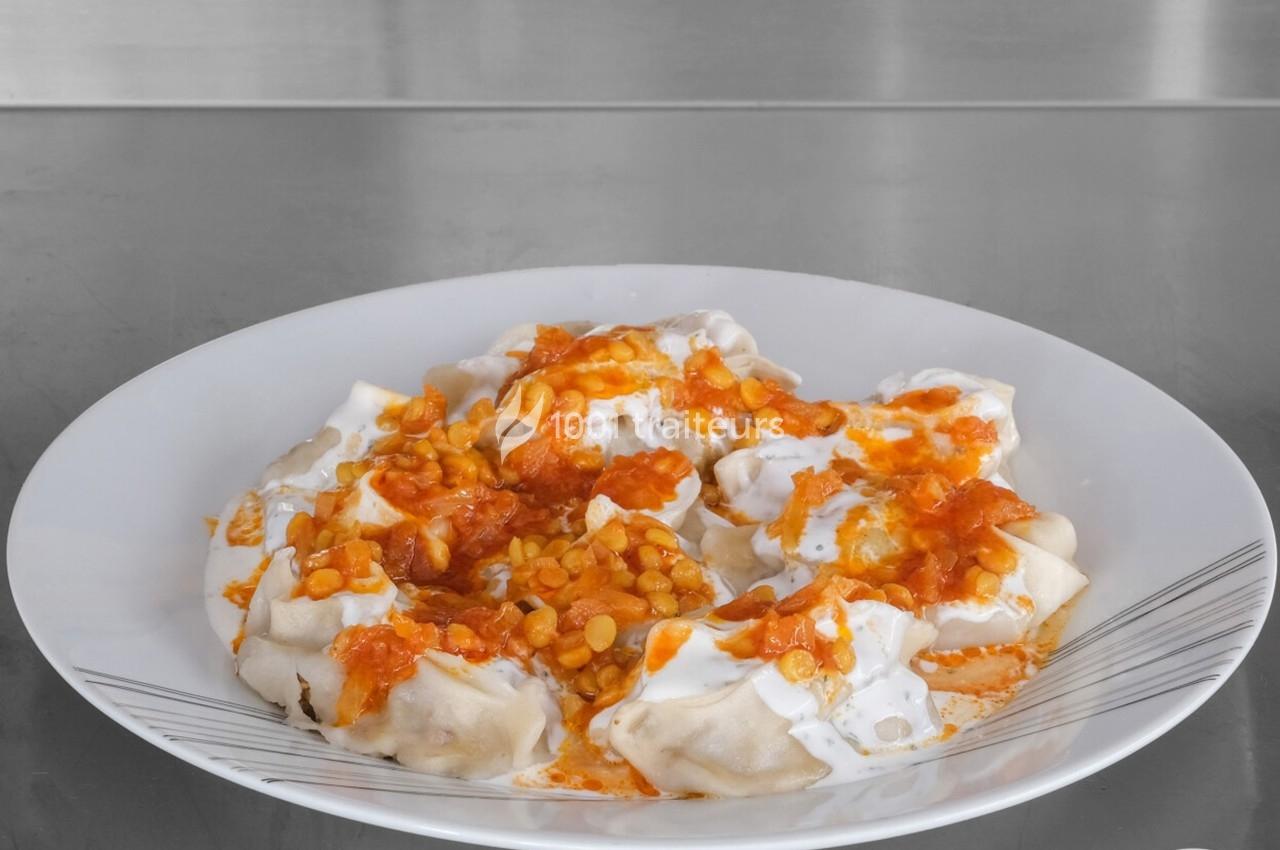 Assiette de raviolis garnis de yaourt, sauce tomate et pois cassés, présentée sur une table en acier inoxydable.