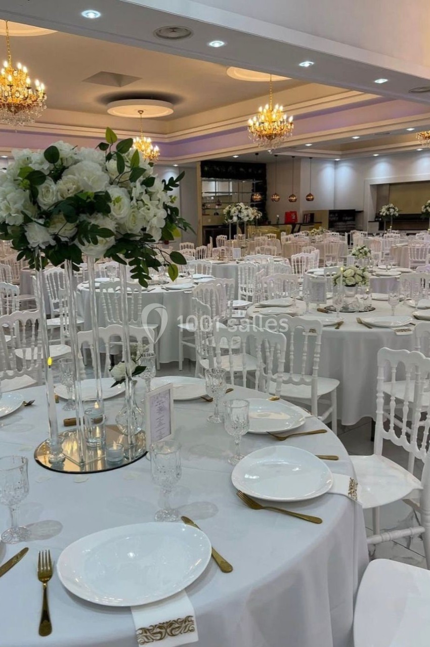 Salle de réception élégante avec tables rondes dressées, chaises blanches et centres de table floraux.