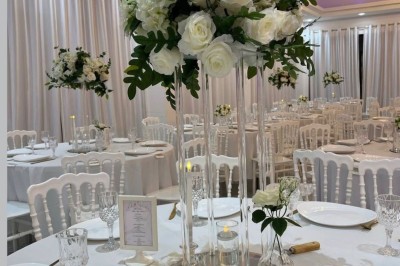 Centre de table avec des roses blanches et feuillage sur un support en verre, dans une salle de réception élégante.