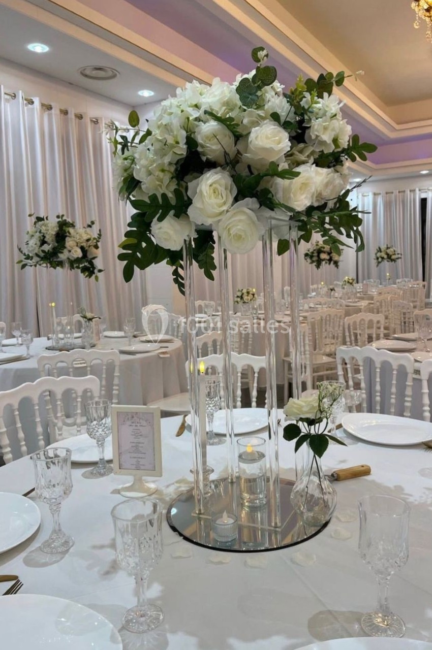 Centre de table avec des roses blanches et feuillage sur un support en verre, dans une salle de réception élégante.