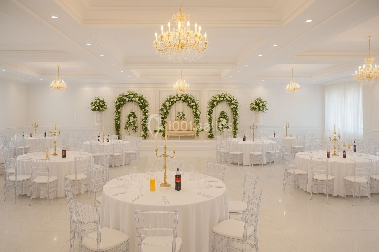 Salle de réception élégante avec tables rondes dressées, chaises transparentes et décorations florales blanches et vertes.