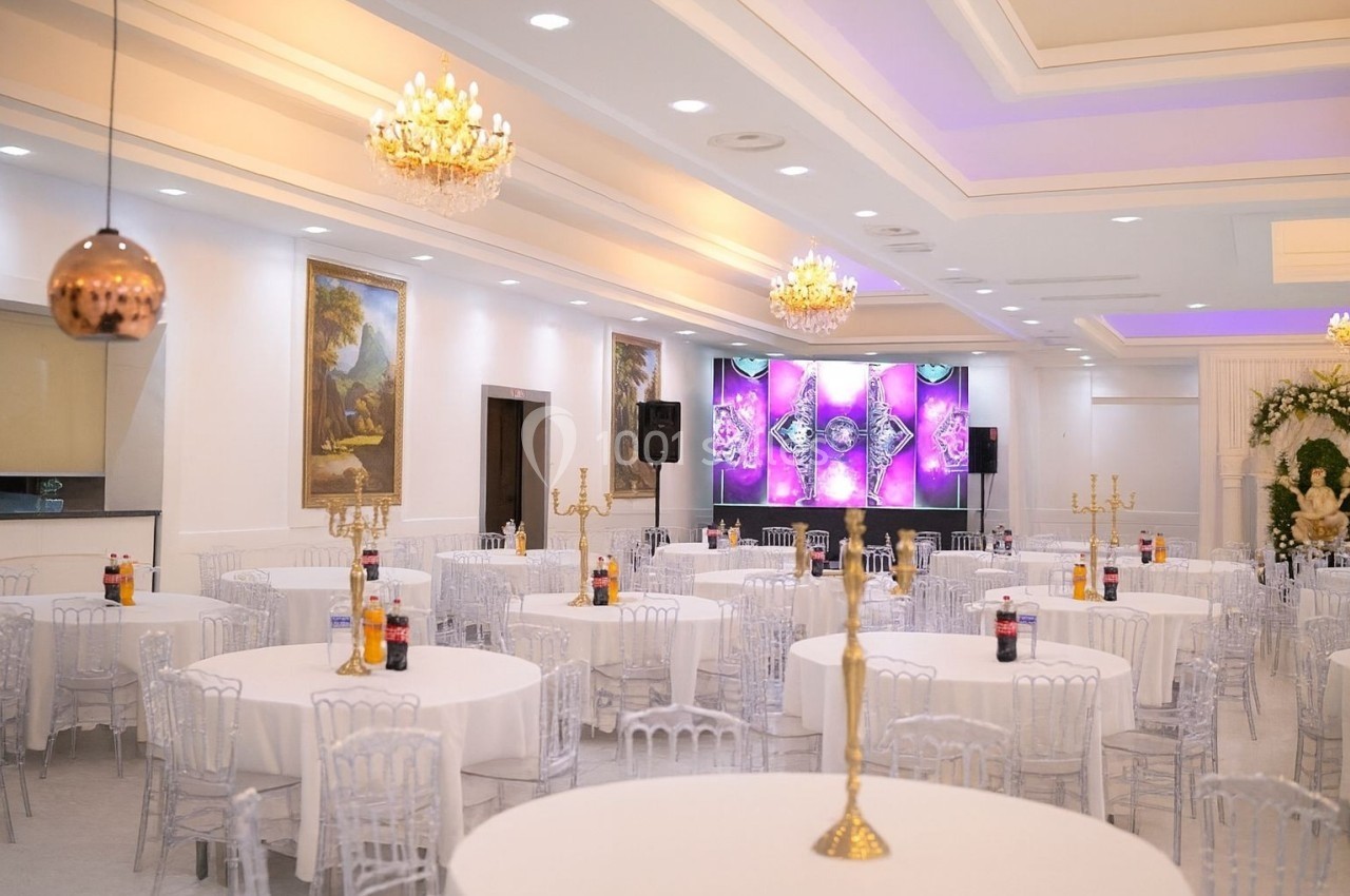 Salle de réception lumineuse avec tables rondes dressées, chandeliers dorés et décorations murales élégantes.