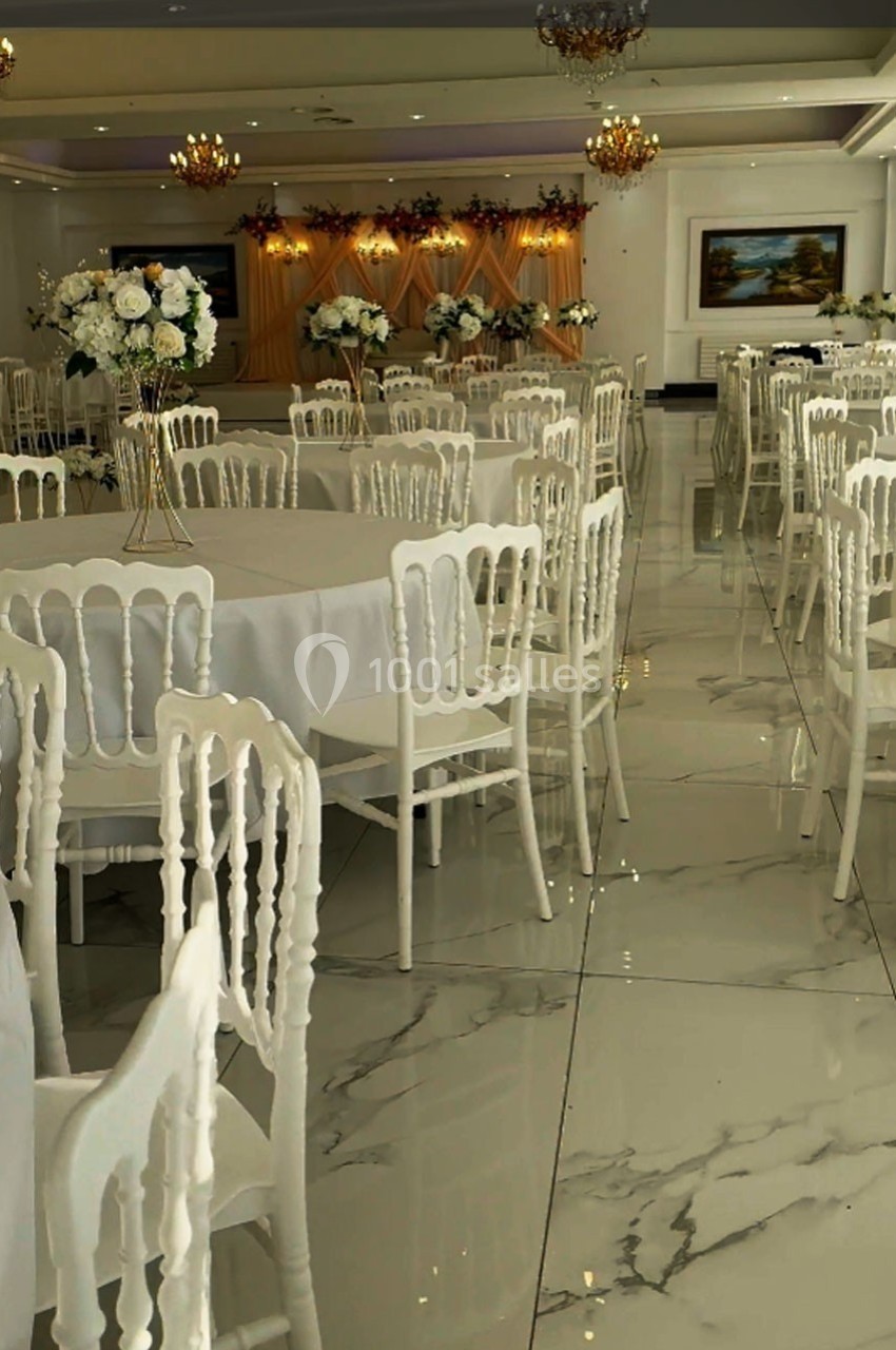 Salle de réception élégante avec tables rondes dressées, chaises blanches et décorations florales.