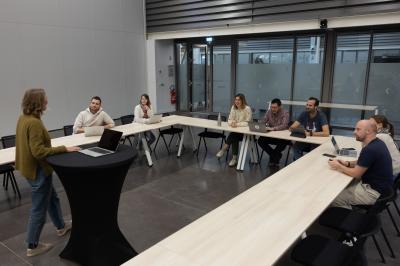 Salle vide avec sol en béton, grandes baies vitrées, murs blancs et plafond industriel avec éclairage suspendu.