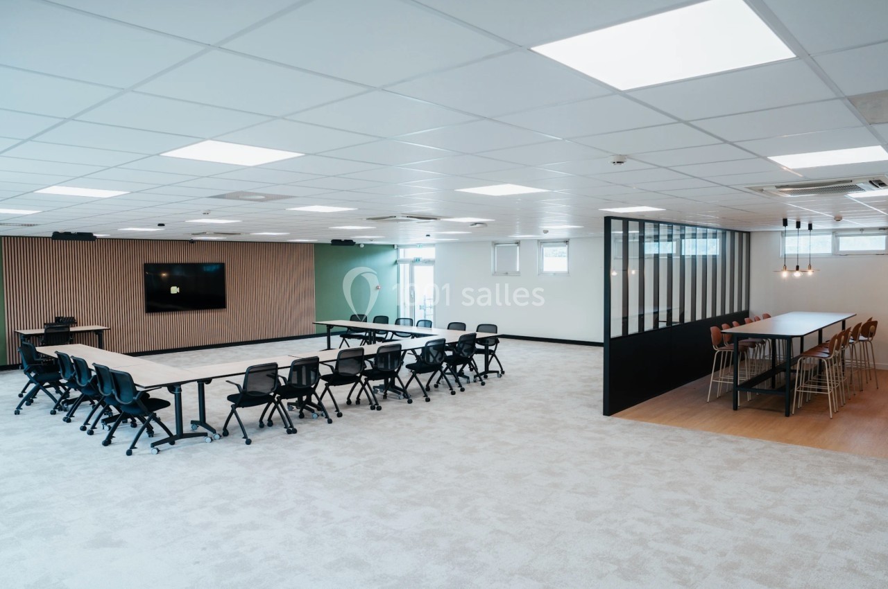 Salle de réunion moderne avec tables, chaises, écran mural et espace de travail adjacent avec table haute.