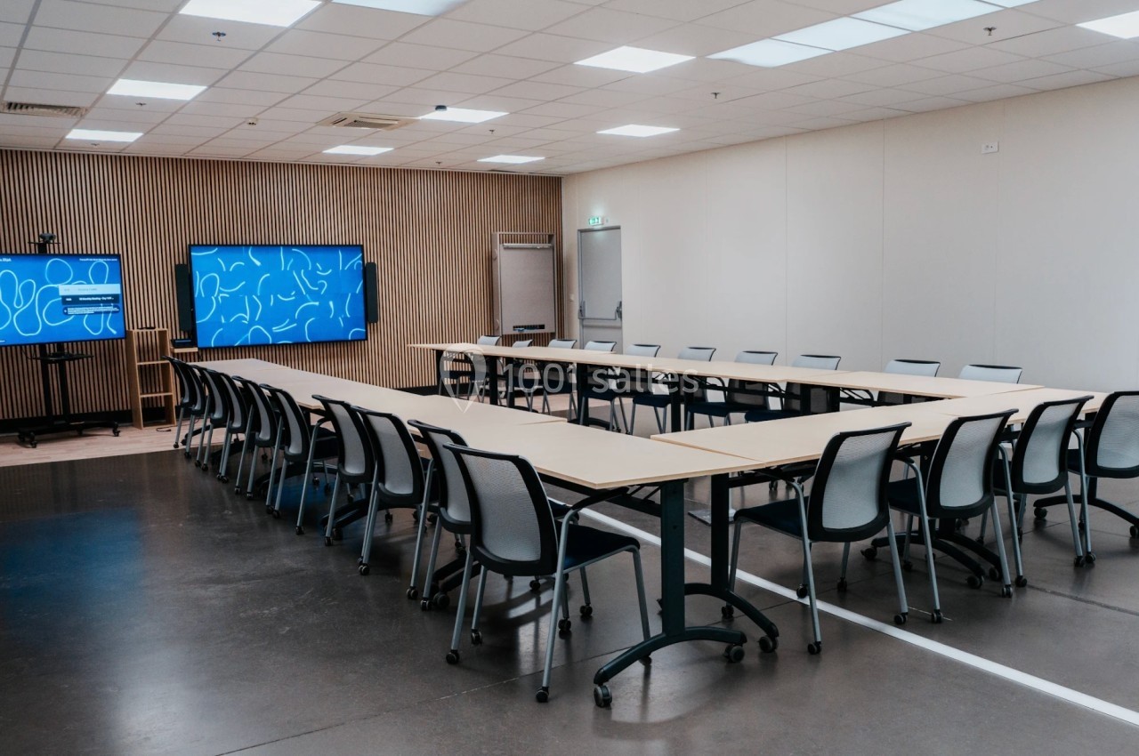 Salle de réunion moderne avec tables disposées en U, chaises noires et écrans interactifs sur les murs.