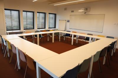 Salle de réunion lumineuse avec tables blanches, chaises orange et un tableau blanc sur pied.
