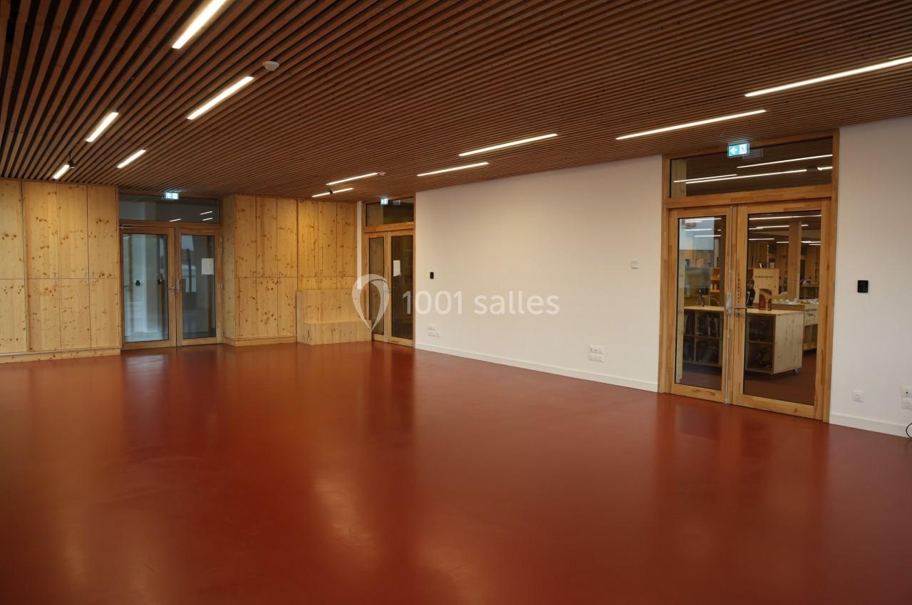 Salle vide avec sol rouge, murs en bois et blanc, plafond en lattes de bois, éclairée par des luminaires encastrés.