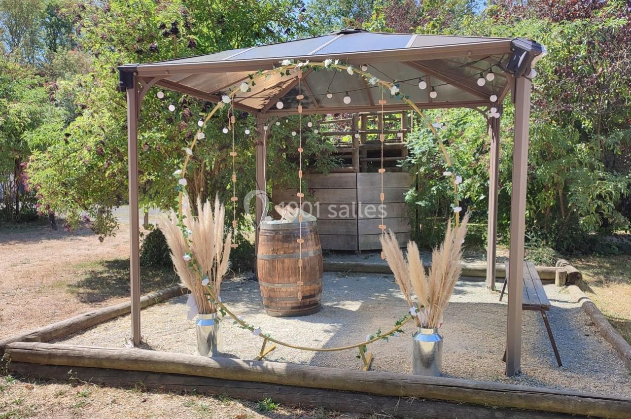 Tonnelle de jardin décorée avec guirlandes lumineuses, cercle orné de fleurs et pampas, et tonneau en bois au centre.