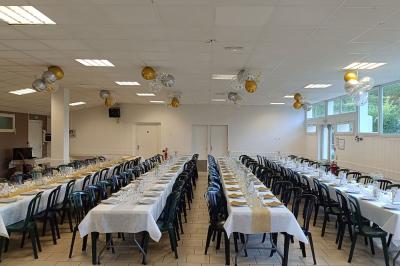 Salon avec téléviseur, fauteuils blancs, chaise orange, tapis à motifs et vue partielle sur une cuisine ouverte.