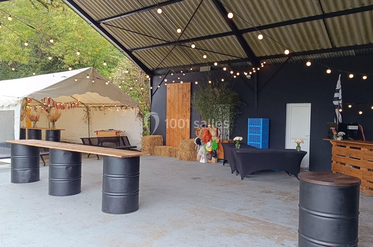 Espace extérieur couvert décoré pour un événement, avec tables en tonneaux, guirlandes lumineuses et tente blanche.