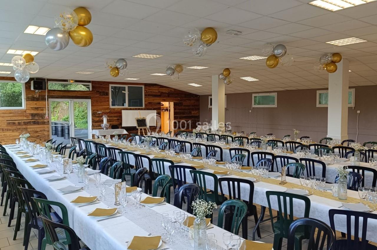 Salle décorée pour un événement avec longues tables dressées, nappes blanches, chemins de table dorés et ballons suspendus.