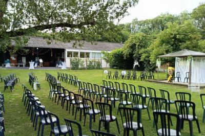 Salon avec téléviseur, fauteuils blancs, chaise orange, tapis à motifs et vue partielle sur une cuisine ouverte.