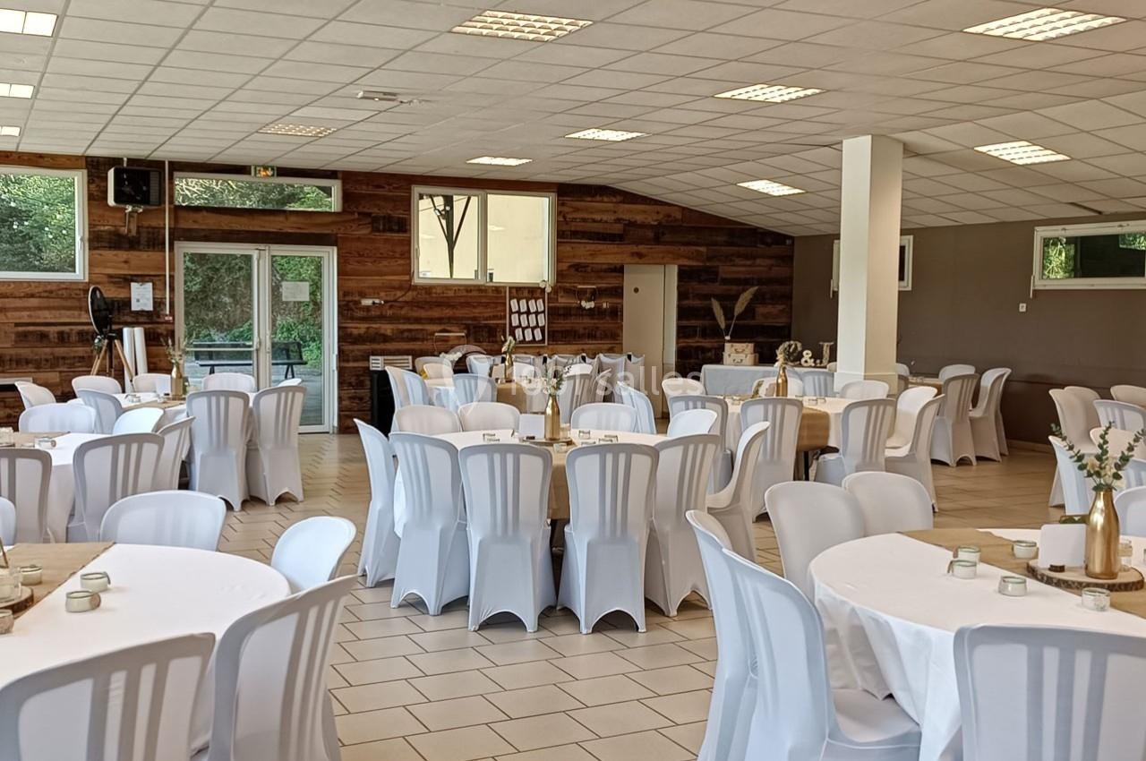 Salle de réception avec des tables rondes dressées de nappes blanches, entourées de chaises en plastique blanc.