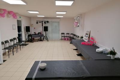 Salon avec téléviseur, fauteuils blancs, chaise orange, tapis à motifs et vue partielle sur une cuisine ouverte.