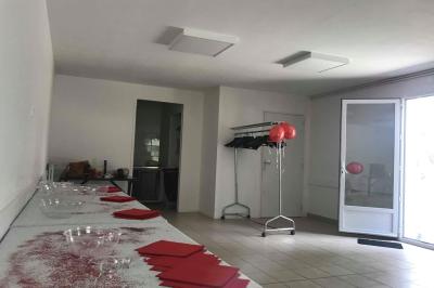 Salon avec téléviseur, fauteuils blancs, chaise orange, tapis à motifs et vue partielle sur une cuisine ouverte.