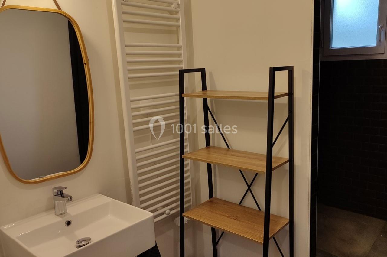 Lavabo avec miroir ovale, étagère en bois, sèche-serviettes mural et fenêtre partiellement visible dans une salle de bain.