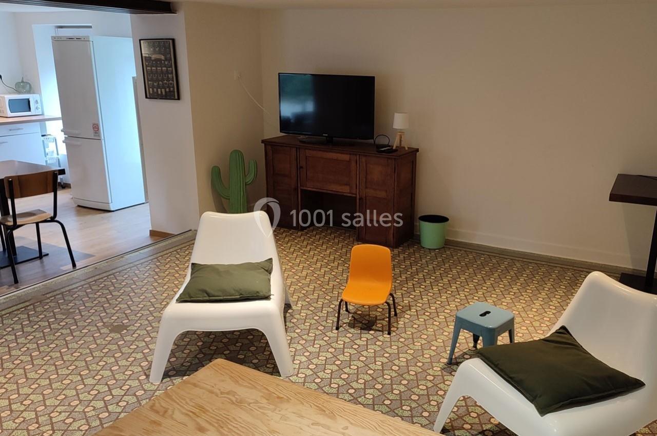 Salon avec téléviseur, fauteuils blancs, chaise orange, tapis à motifs et vue partielle sur une cuisine ouverte.