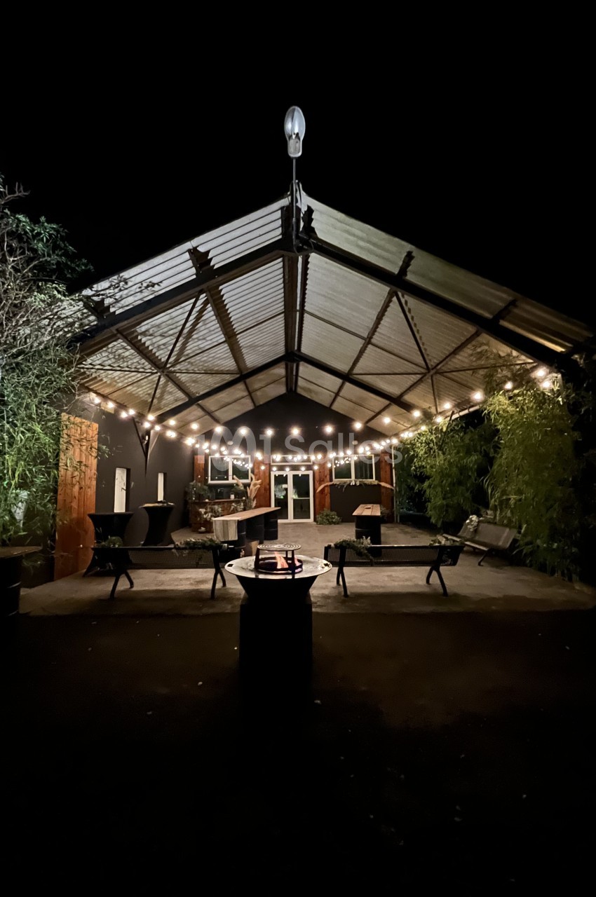 Terrasse couverte de nuit éclairée par des guirlandes de guinguettes, avec tables hautes, bancs métalliques et brasero central. Wait, I should avoid "guinguettes" as it's an interpretation; use "guirlandes lumineuses". Also count characters: must be