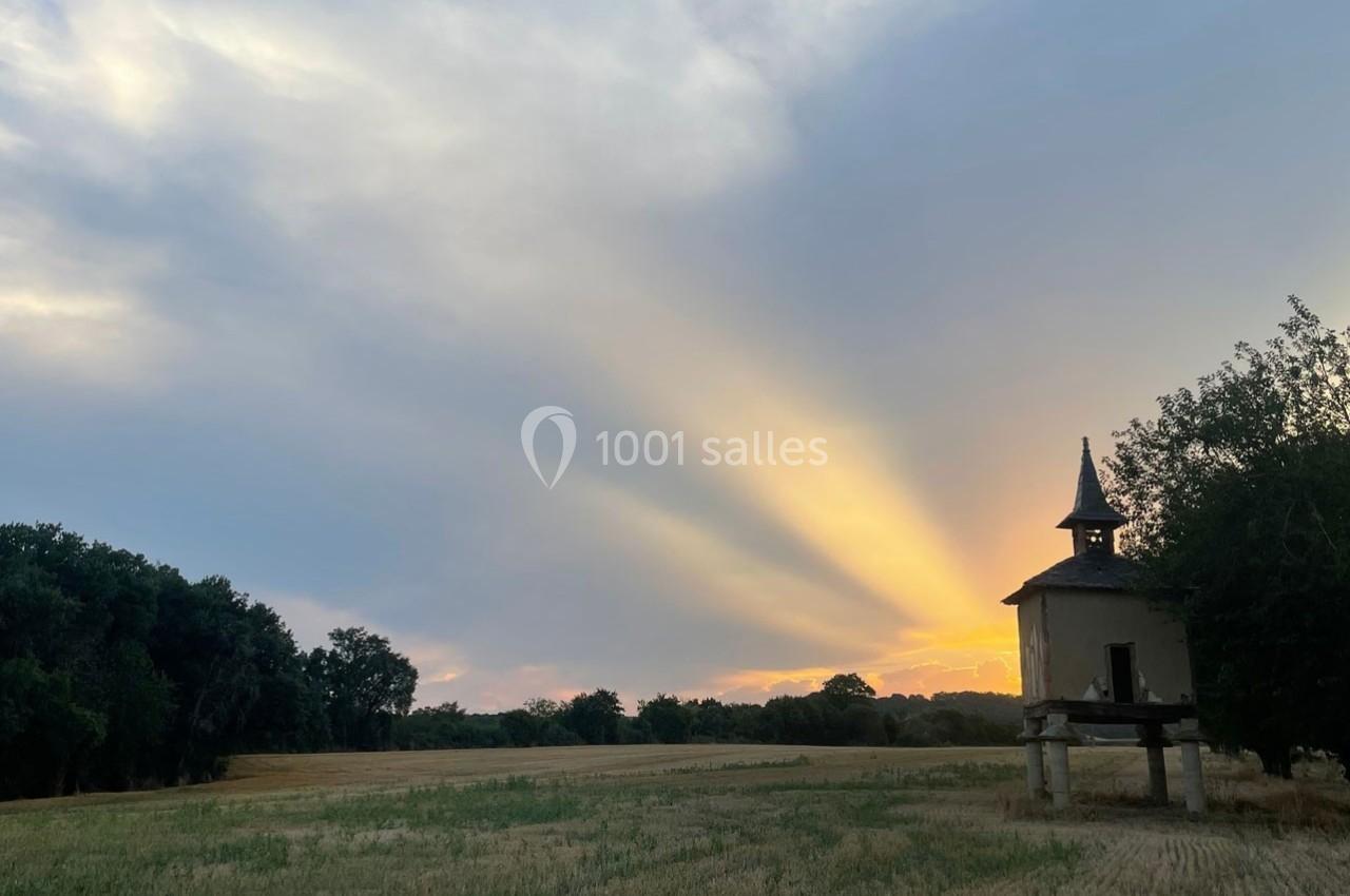 Rayons de soleil traversant un ciel nuageux au crépuscule, éclairant un champ et un pigeonnier en pierre.