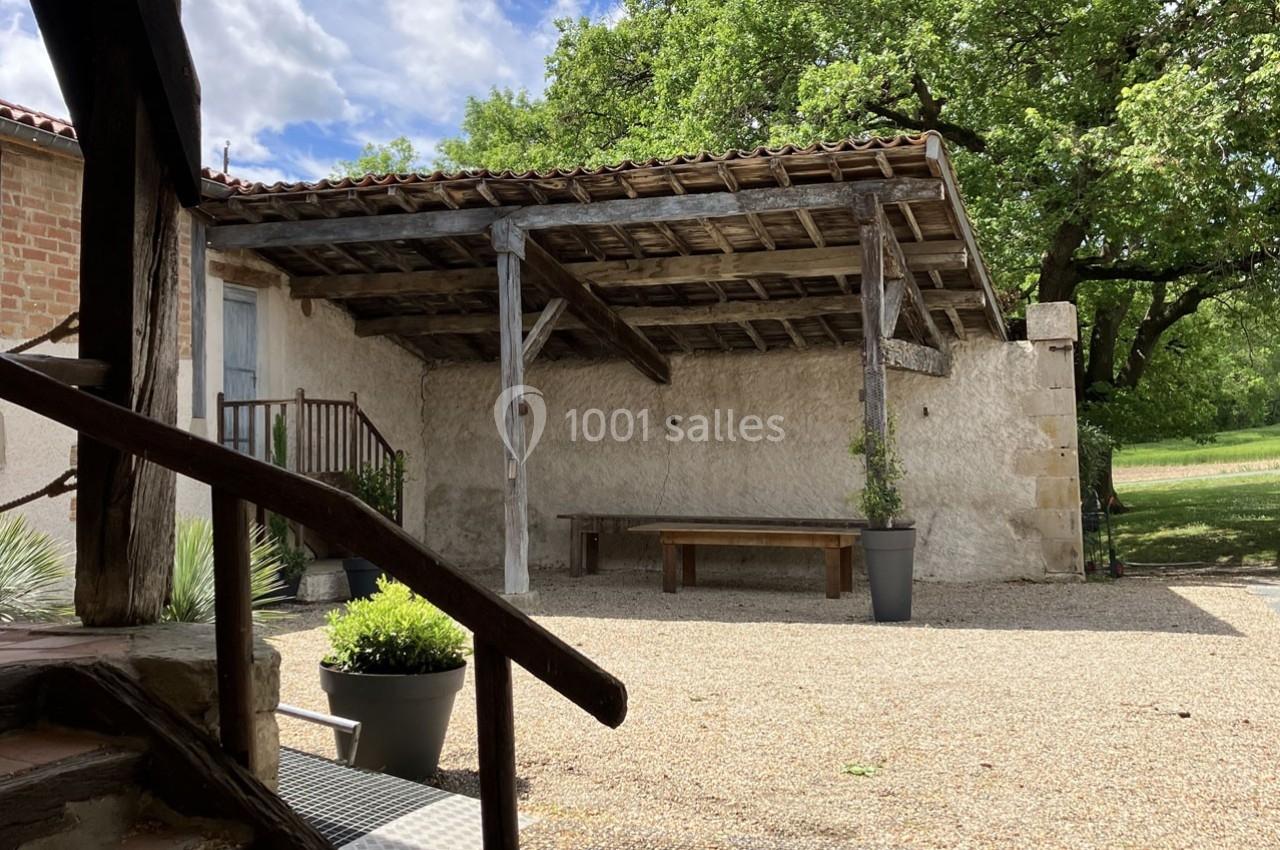 Cour extérieure avec auvent en bois, banc en bois, pots de fleurs et arbres en arrière-plan sous un ciel partiellement…
