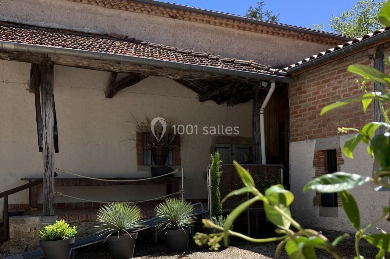 Façade d'une maison en pierre avec toit en tuiles, plantes en pots et hamac dans une cour ensoleillée.