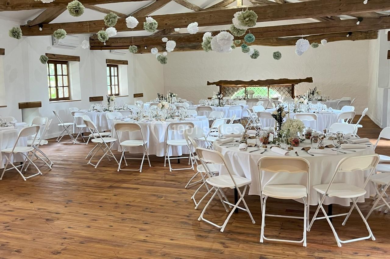 Salle de réception avec tables rondes dressées, chaises blanches et décorations suspendues sous un plafond en poutres…