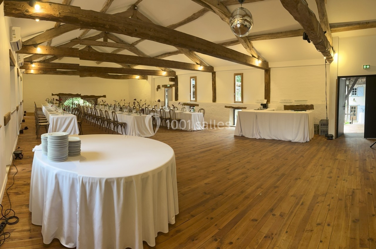 Salle de réception avec poutres apparentes, tables dressées et nappées, parquet en bois et lumière naturelle.