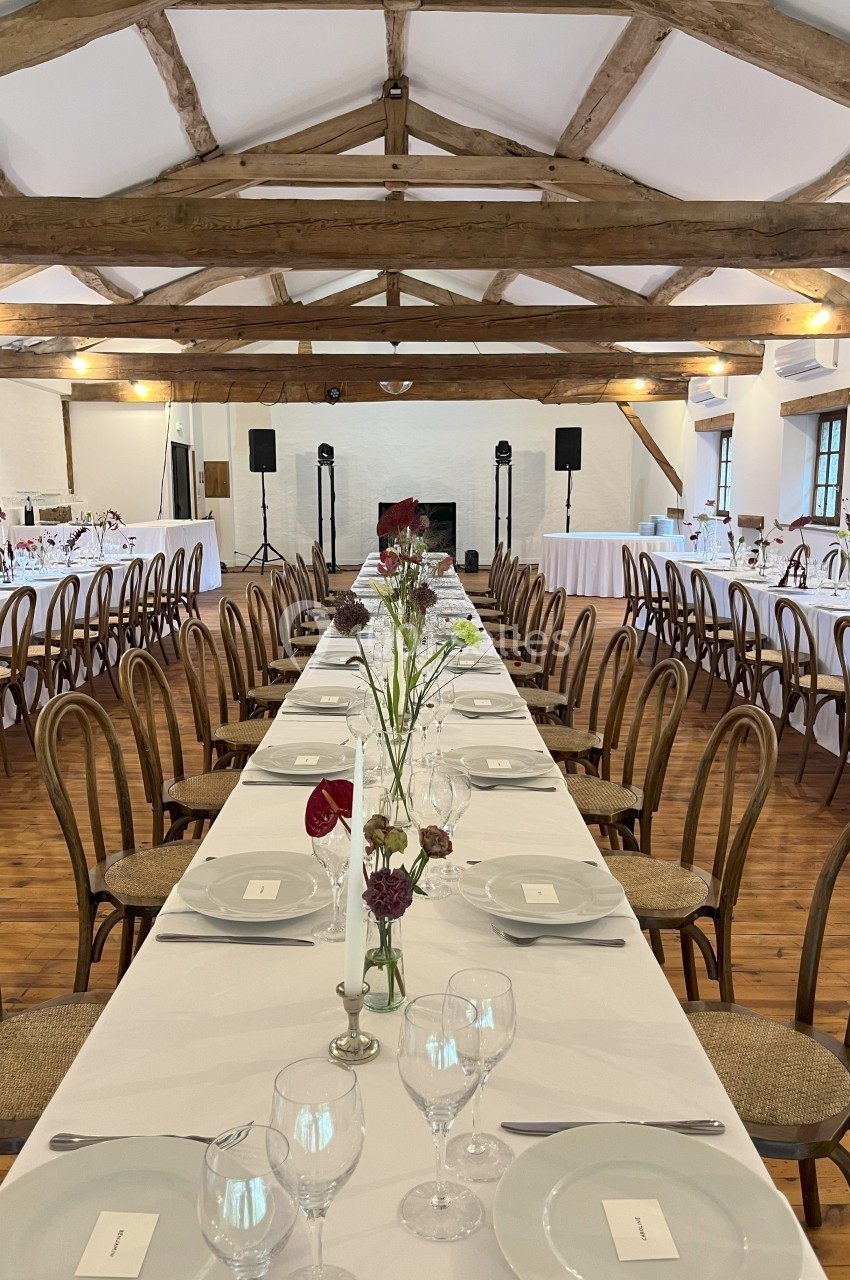Grande salle de réception avec tables dressées, chaises en bois, poutres apparentes et décoration florale simple.
