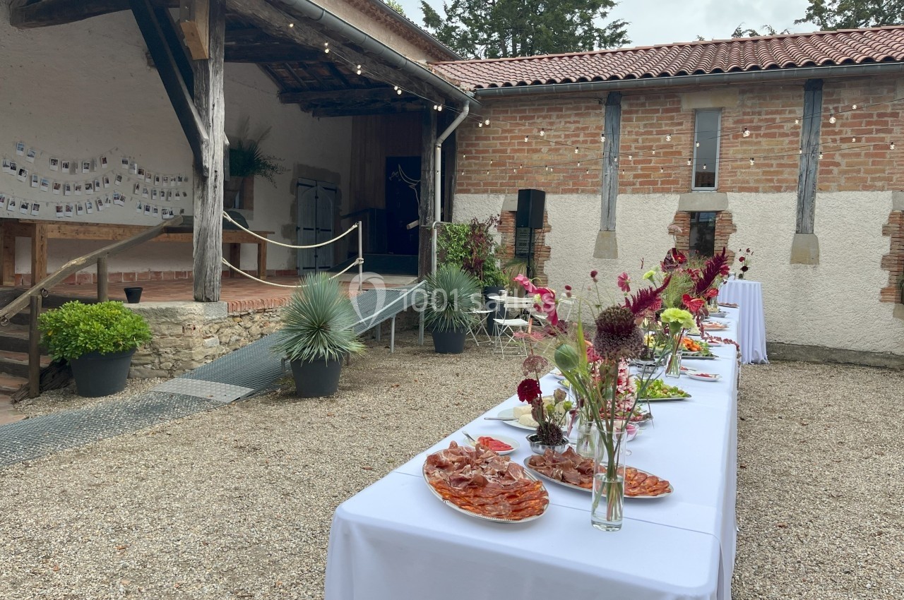 Table dressée avec des plats de charcuterie et fleurs, dans une cour extérieure avec bâtiment en briques et bois.