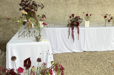 Salle rustique avec murs en pierre, poutres apparentes, table dressée, chaises en bois et coin salon avec canapé.
