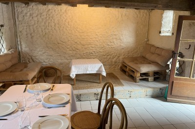 Salle rustique avec murs en pierre, poutres apparentes, table dressée, chaises en bois et coin salon avec canapé.