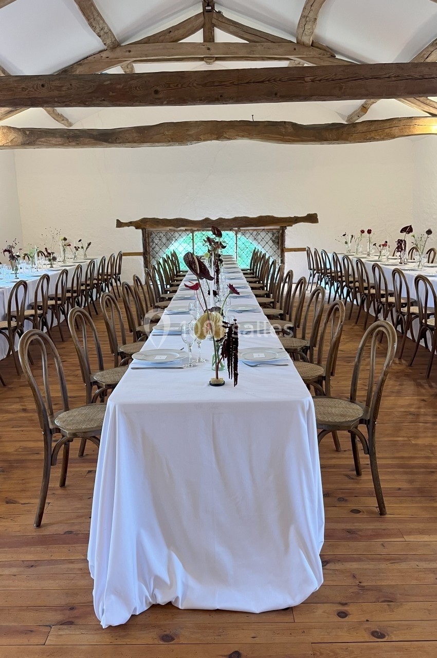 Salle de réception avec tables alignées, nappes blanches, chaises en bois et décorations florales minimalistes.