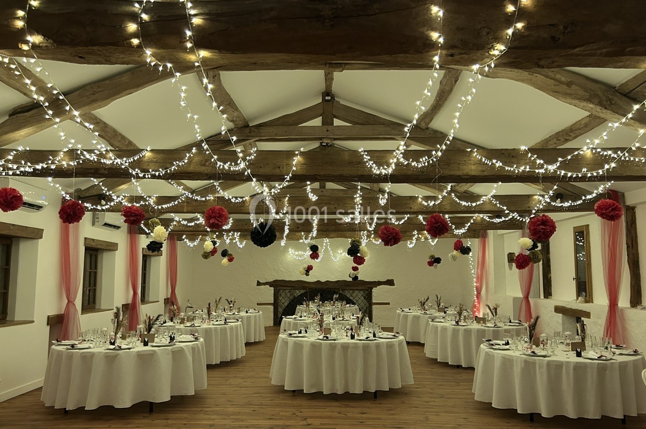 Salle de réception décorée avec des guirlandes lumineuses, des tables rondes dressées et des fleurs suspendues.