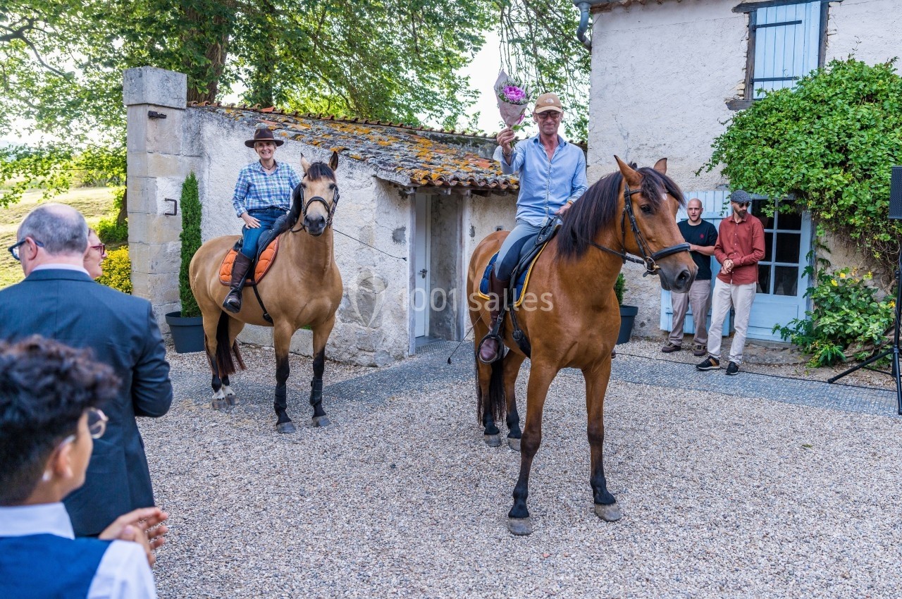 Deux cavaliers sur des chevaux devant une maison en pierre, observés par un petit groupe de personnes.