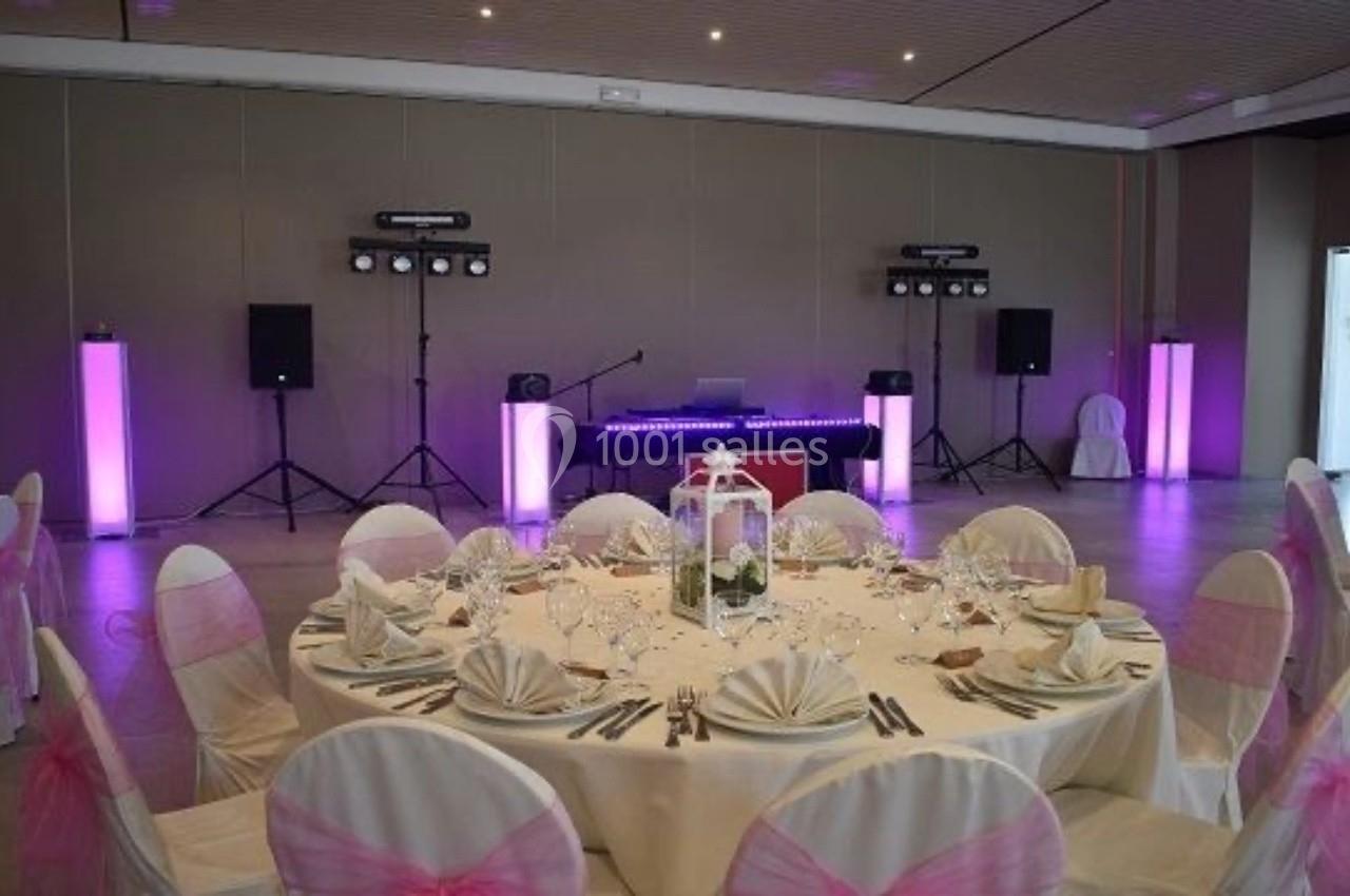 Salle décorée pour un événement avec table ronde dressée, chaises ornées de nœuds roses et équipement de sonorisation en…