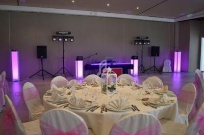 Salle de réception décorée avec une table ronde dressée, chaises blanches ornées de nœuds roses et éclairage lumineux.