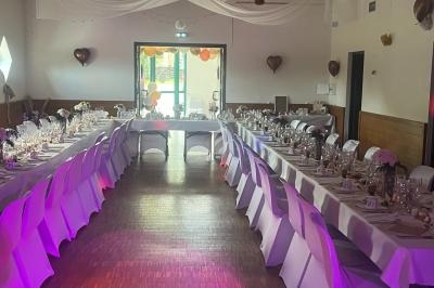Salle décorée pour un événement avec tables alignées, nappes blanches, vaisselle élégante et ballons en arrière-plan.
