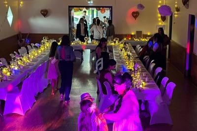 Salle décorée pour une réception avec longues tables illuminées, invités discutant et enfants jouant au premier plan.