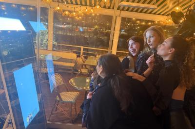 Deux femmes souriantes se tiennent près d'une borne photo dans un espace intérieur avec des tables et des chaises en arrière…