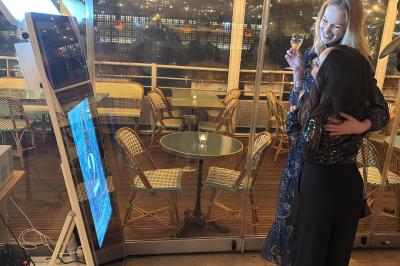 Deux femmes souriantes se tiennent près d'une borne photo dans un espace intérieur avec des tables et des chaises en arrière…