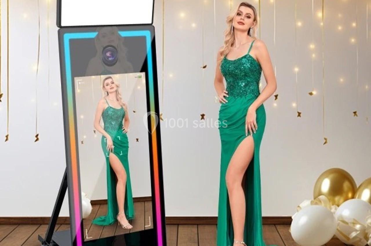 Une femme en robe verte élégante pose devant un miroir interactif, entourée de ballons et de décorations dorées.