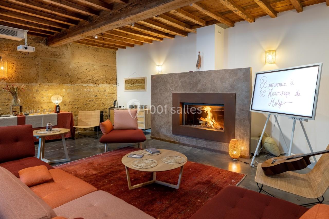 Salon chaleureux avec cheminée allumée, canapé rouge, tables basses en bois et écran affichant un message de bienvenue.