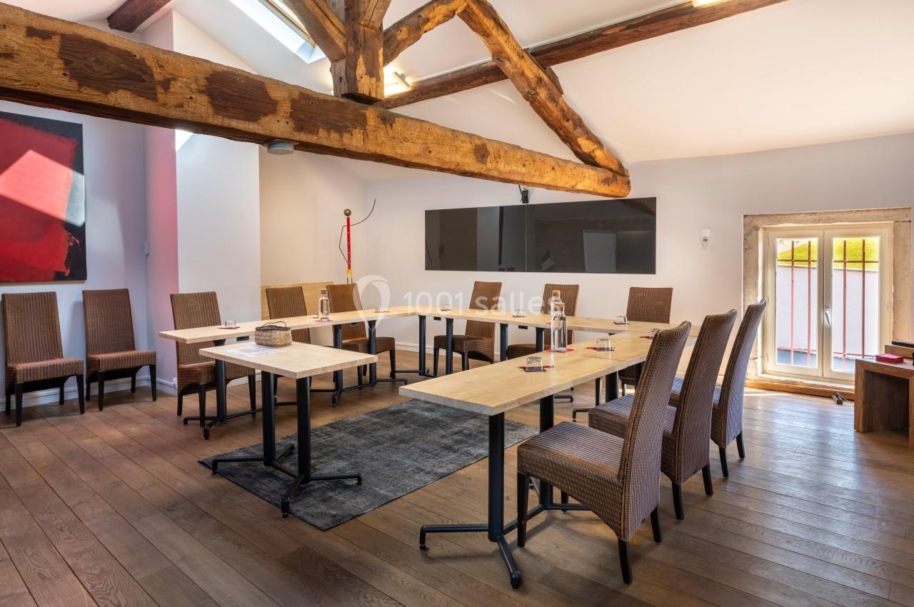 Salle de réunion lumineuse avec poutres apparentes, tables en U, chaises en osier et écran mural.