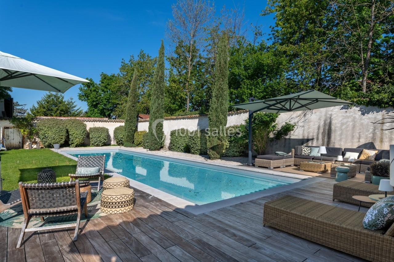 Piscine extérieure entourée de végétation, avec terrasse en bois, chaises longues et parasols sous un ciel bleu.
