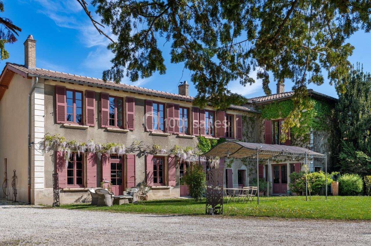 Grande maison en pierre avec volets rouges, pergola ombragée et jardin arboré sous un ciel bleu.